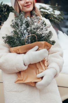 Woman in white hat and winter fur coat with pine branch in city, lights and decorations on background.