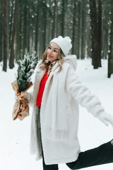 Young blonde woman wears white fur coat, knitted hat and mittens, posing with fir tree branches wrapped with craft paper in winter forest. Winter fashion