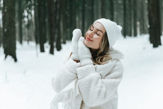 Young woman Enjoying snowy winter forest and warm clothes. Dressed white coat scarf hat and mittens fold palms to face.