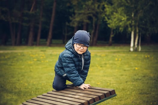 A little boy with glasses plays on a wooden chaise longue in the park during the day