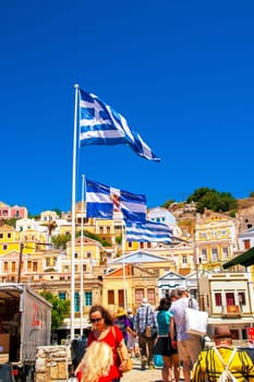 SYMI, Greece - June 03, 2021. Tuorists at the harbour of Symi Town. Colorful villas stretch up the steep slopes.