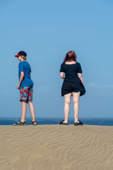 Two teenagers stand on a sand dune, with sea and blue sky