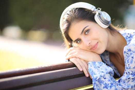 Relaxed woman looks at you listening audio with headphone in a park