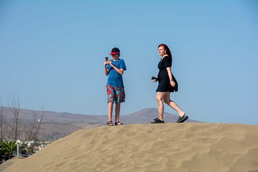 Two teenagers are standing on a sand dune with their cell phones, the boy is looking at his smartphone
