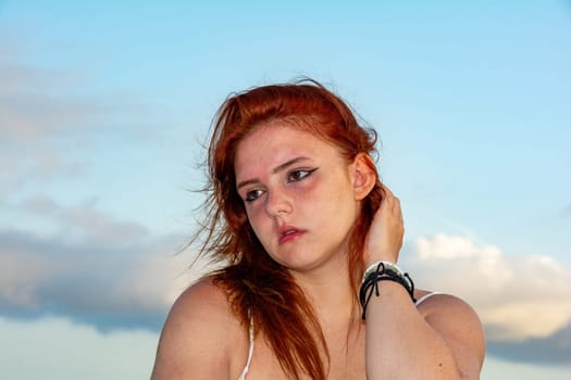 Portrait of a young girl with red hair, blue sky and clouds, hair tousled by the wind