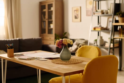 Wooden table with papers, crayons and grey vase with flowers standing in front of camera in the center of spacious living room