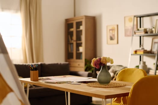 Wooden table with flowers in vase, papers and crayons standing in the center of spacious living room of modern apartment