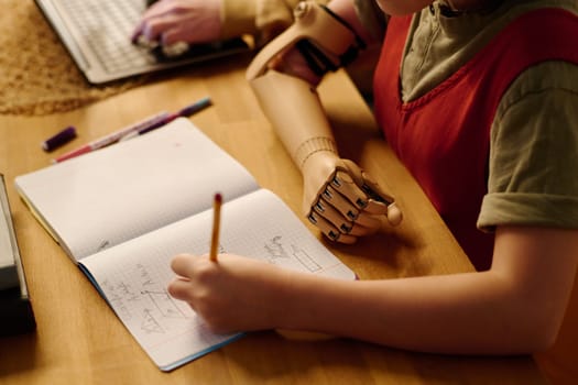 Hand prosthesis of youthful diligent schoolgirl making notes in copybook while sitting by desk and doing homework on geometry