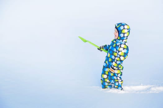 Laughing a little boy in winter with a green shovel in overalls makes his way through a snow-covered field