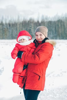 Mom and daughter in red jumpsuits are standing in a field next to a forest in winter for a winter photo shoot