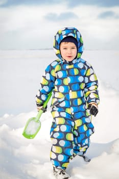 Laughing a little boy in winter with a green shovel in overalls makes his way through a snow-covered field