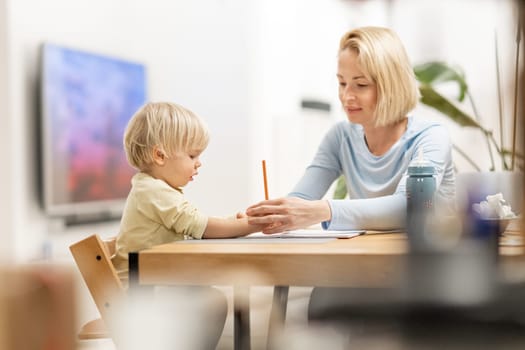 Caring young Caucasian mother and small son drawing painting in notebook at home together. Loving mom or nanny having fun learning and playing with her little 1,5 year old infant baby boy child