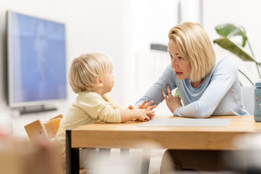 Caring young Caucasian mother and small son drawing painting in notebook at home together. Loving mom or nanny having fun learning and playing with her little 1,5 year old infant baby boy child