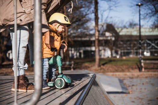 Father supervises his fearless small toddler boy while riding baby scooter outdoors in urban skate park. Child wearing yellow protective helmet.