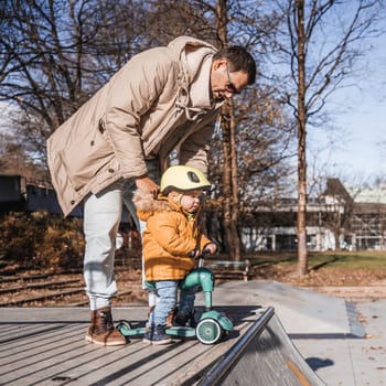 Father supervises his fearless small toddler boy while riding baby scooter outdoors in urban skate park. Child wearing yellow protective helmet.
