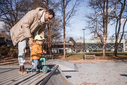 Father supervises his fearless small toddler boy while riding baby scooter outdoors in urban skate park. Child wearing yellow protective helmet.