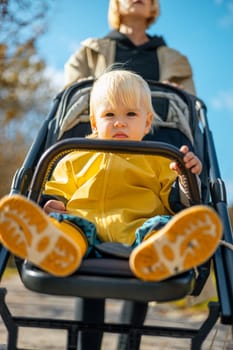 Little toddler child wearing yellow rain coat and rain boots being pushed in stroller by her mother in city park on a sunny autumn day after the rains were gone