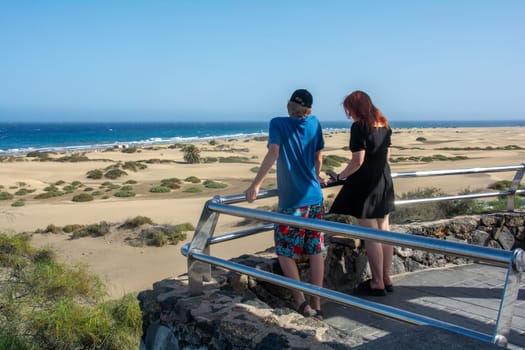 Two teenagers stand on a viewing platform on the sandy beach and look at the dunes of Maspalomas on Gran Canaria, Spain
