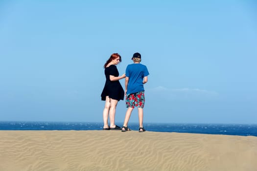 Two teenagers stand on a sand dune, with sea and blue sky