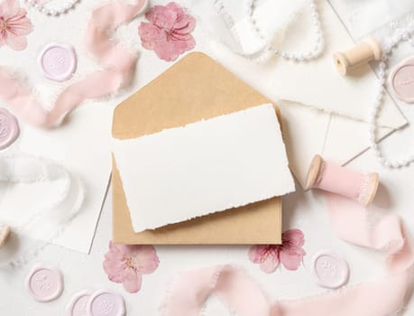 Card and envelope near pink decorations, seals and silk ribbons on white table top view, wedding mockup. Romantic scene with horizontal blank card flat lay. Valentines, Spring or Mothers day concept