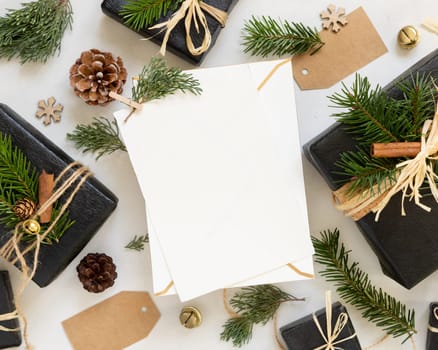 Paper card between Christmas decor, black gift boxes and green fir branches top view. Vertical blank card mockup, flat lay, copy space. Christmas or New Year Holiday greeting card