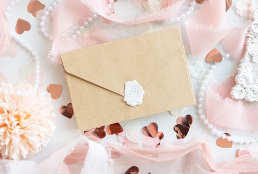Sealed envelope near pink decorations, hearts and silk ribbons on white table top view, mockup. Romantic scene with vertical blank card flat lay. Wedding, Valentines, Spring or Mothers day stationery