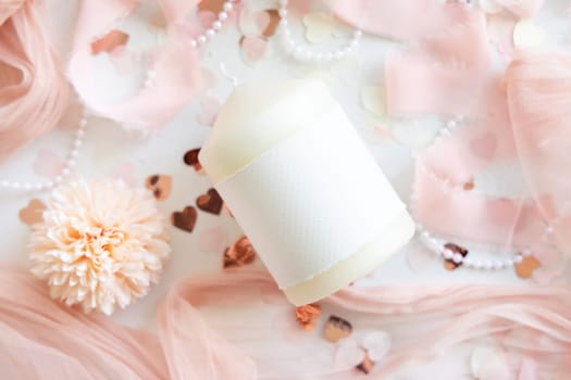 Candle with blank label near pink decorations, hearts and silk ribbons on white table top view. Romantic home decor. brand packaging mock up. Home decoration, romantic interior