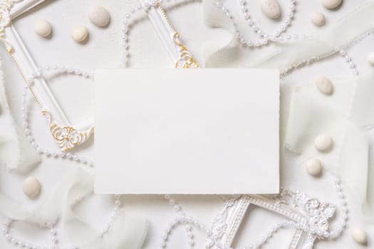 Blank paper card near white frames, pearls, pebbles and silk ribbons on white table top view, wedding mockup. Romantic scene with horizontal card flat lay. Valentines, Spring or Mothers day concept