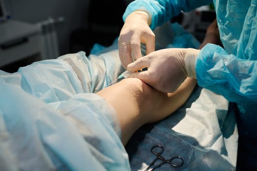 Gloved hands of professional surgeon holding syringe with anaesthesia injection by knee of young female patient on operation table