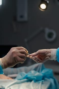 Gloved hand of assistant passing sterile scalpel or some other surgical tool against patient covered with napkin during operation