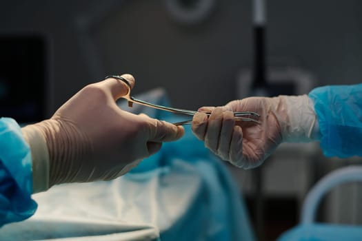 Gloved hands of assistant and surgeon passing and taking steel medical tool during surgical operation against patient covered with napkin