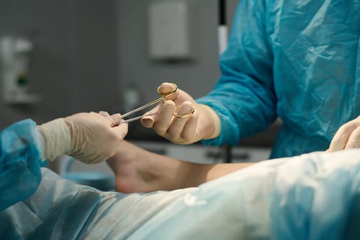 Gloved hand of assistant passing steel sterile forceps to surgeon during surgical operation over patient covered with napkin