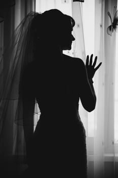 The black silhouette of the bride standing at the window into the room in close-up during the day