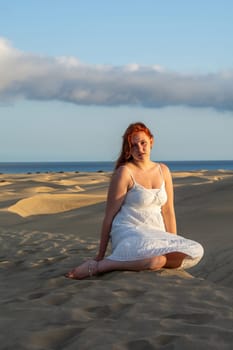 A young pretty red-haired girl in a white dress sits in the sand dunes in the late afternoon and enjoys the last rays of sunshine
