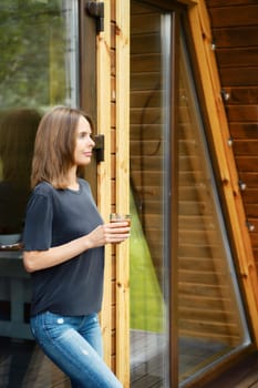 Cute woman came out of a wooden house, leaned against a glass door and drinks juice from a glass