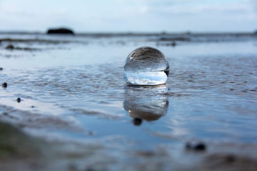 A glass ball lies in the waves on the sandy beach, the sea and the setting sun are reflected in the ball