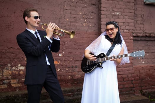 The groom plays the trumpet and the bride plays the guitar at a wedding photo shoot against a brick wall on the street