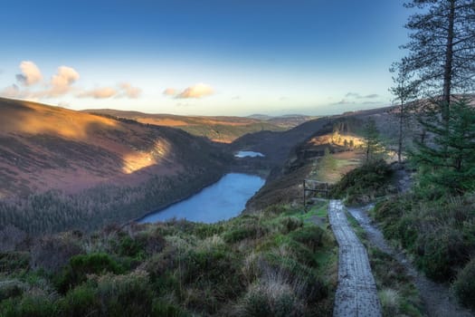 Wooden boardwalk on the top of a mountain with scenic view on Glendalough lakes and forest. Hiking at sunset in Wicklow Mountains, Ireland