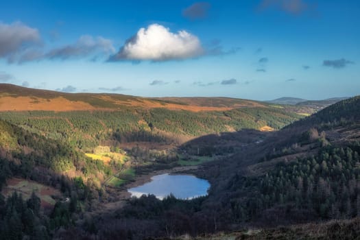 Glendalough lake and cemetery in the valley. Forest on the hills illuminated by sunlight. Hiking in beautiful autumn Wicklow Mountains, Ireland