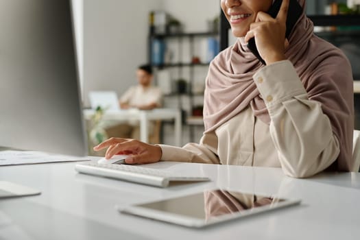 Cropped shot of young smiling office manager in hijab talking on mobile phone and clicking mouse while sitting in front of computer