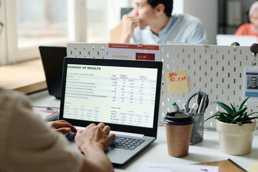 Financial data on screen of laptop being used by young male economist sitting in front of pensive colleague looking through office window