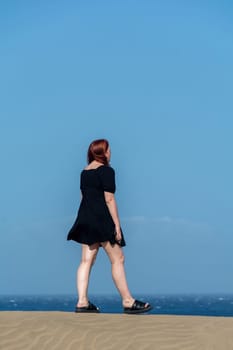 Young girl  in a black  short summer dress stands on a sand dune with a blue sky
