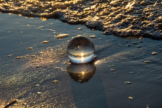 A glass ball lies in the waves on the sandy beach, the sea and the setting sun are reflected in the ball