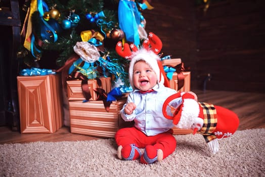 Laughing a little boy with deer antlers on his head is sitting in the studio near the Christmas tree and a bunch of gifts
