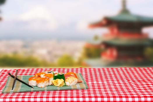 Table top on sushi background. Close-up of various kinds of sushi and chopsticks served on green bamboo mat on a red checkered tablecloth over abstract blurred japanese landscape.
