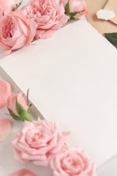 Blank card near light pink roses and petals on white table close up, wedding mockup, copy space. Romantic scene with vertical card and pastel decor
