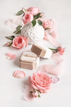 Engagement ring in a cream velvet box near light pink roses, buds and silk ribbons close up, romantic wedding preparations. Marriage proposal concept, valentines day scene