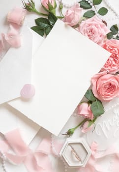 Blank card and envelope near light pink roses, engagement ring in a box and silk ribbons on white table top view, wedding mockup. Romantic flat lay with vertical card and pastel decor