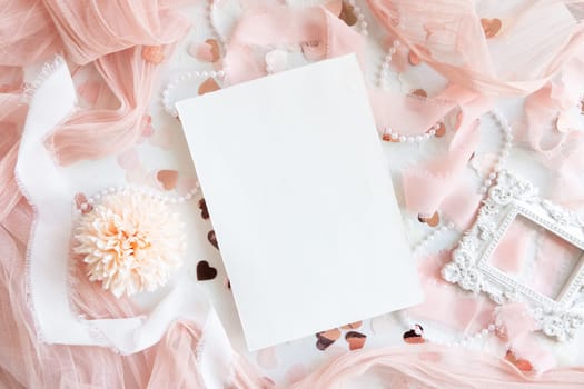 Blank paper card near pink decorations, hearts and silk ribbons on white table top view, mockup. Romantic scene with vertical blank card flat lay. Wedding, Valentines, Spring or Mothers day stationery
