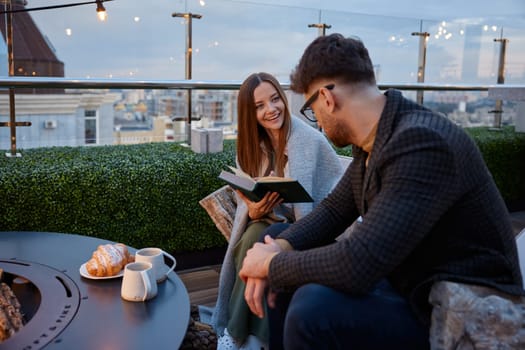 Young girlfriend reading for boyfriend while couple resting at roof top cafe. Excited female sharing impression of favorite book. Young family leisure activity in evening